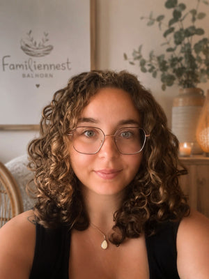 Lächelnde Hebamme mit lockigem Haar und Brille vor dem Logo des Familiennest Balhorn – Expertin für Neugeborenenversorgung