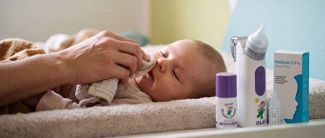 Mutter befeuchtet die verstopfte Nase ihres Babys mit Kochsalzlösung, daneben Nasensauger, Rüsselcreme auch die Pädia Kochsalzlösung auf dem Wickeltisch